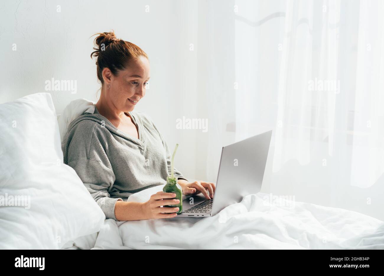 Una donna in casa abiti casual a letto con un computer portatile e un cocktail detox sta lavorando o shopping. Foto Stock