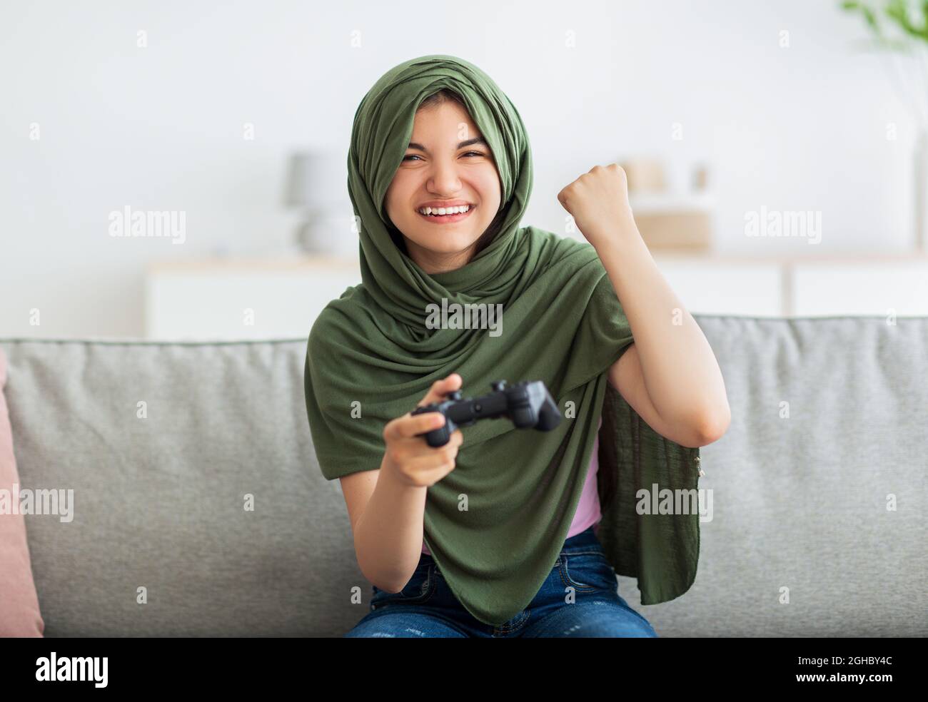 Ragazza teen indiana trionfante in hijab con joystick che gioca a videogame, facendo sì gesto, celebrando la vittoria a casa Foto Stock