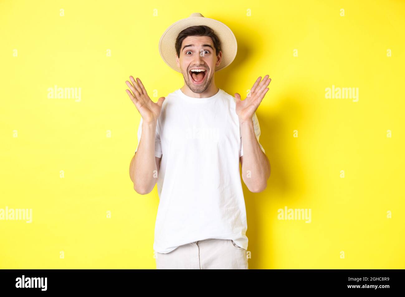Concetto di turismo ed estate. Felice giovane uomo in cappello di paglia che guarda stupito, reagendo a sorpresa, in piedi su sfondo giallo Foto Stock