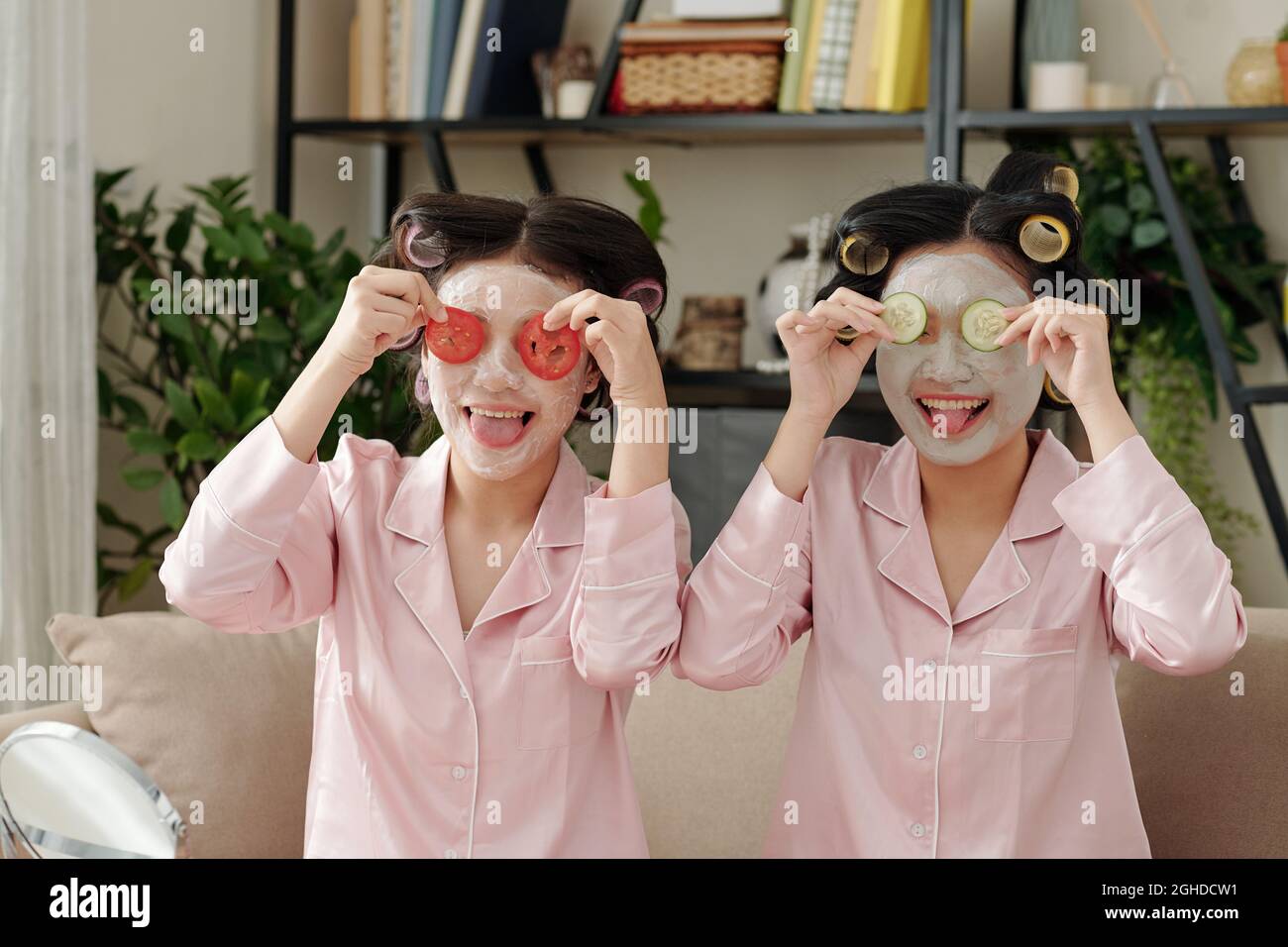 Donne giovani piuttosto felici con maschera di argilla sulla creazione di volti divertenti con fette di pomodoro e cetriolo e mettendo lingua fuori Foto Stock