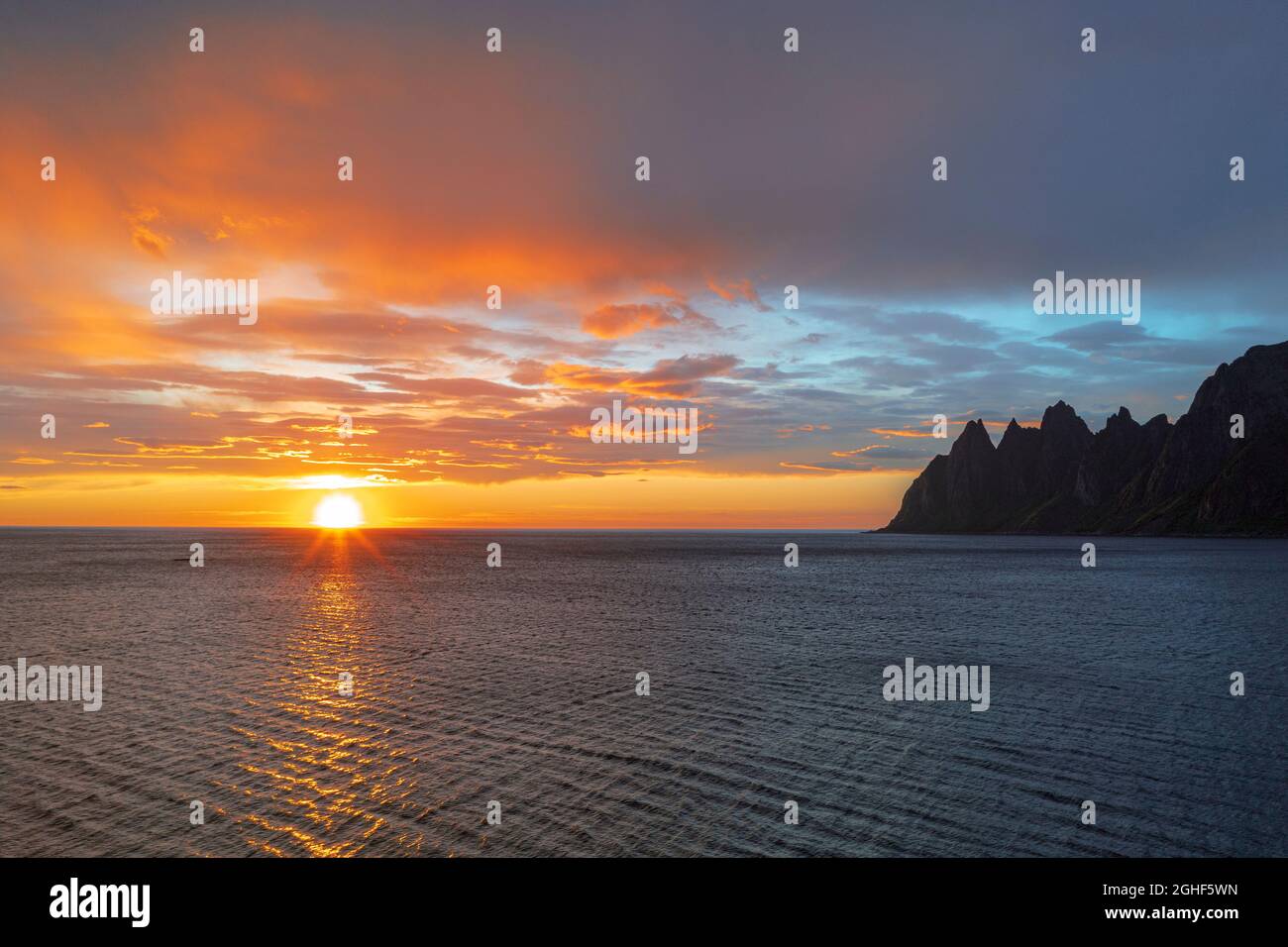 Il sole di mezzanotte si riflette nel mare con le cime dei denti del Diavolo sullo sfondo, Tungeneset, Senja, contea di Troms, Norvegia Foto Stock