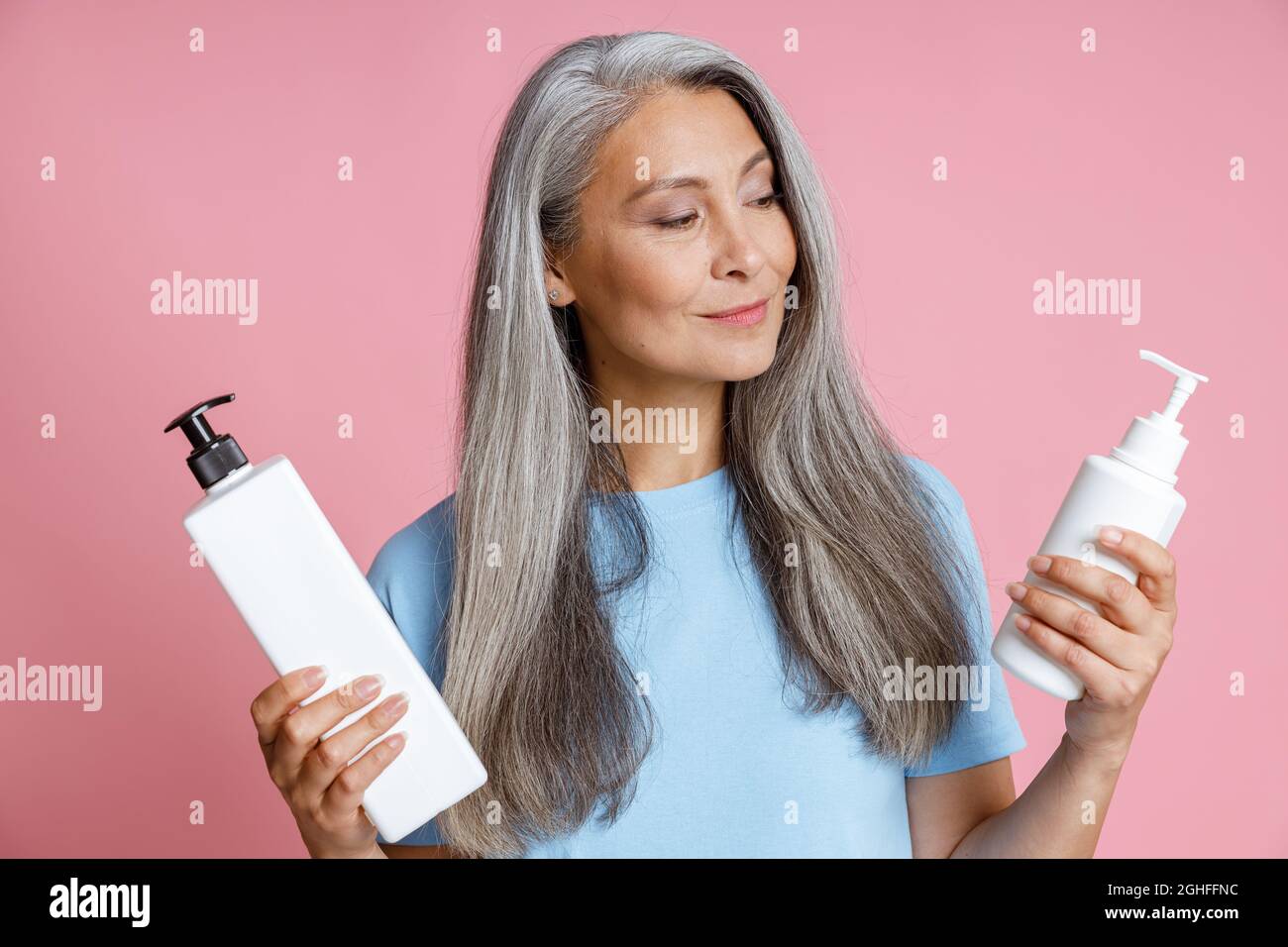 Positivo donna asiatica matura ichooses miglior prodotto per la cura dei capelli su sfondo rosa Foto Stock