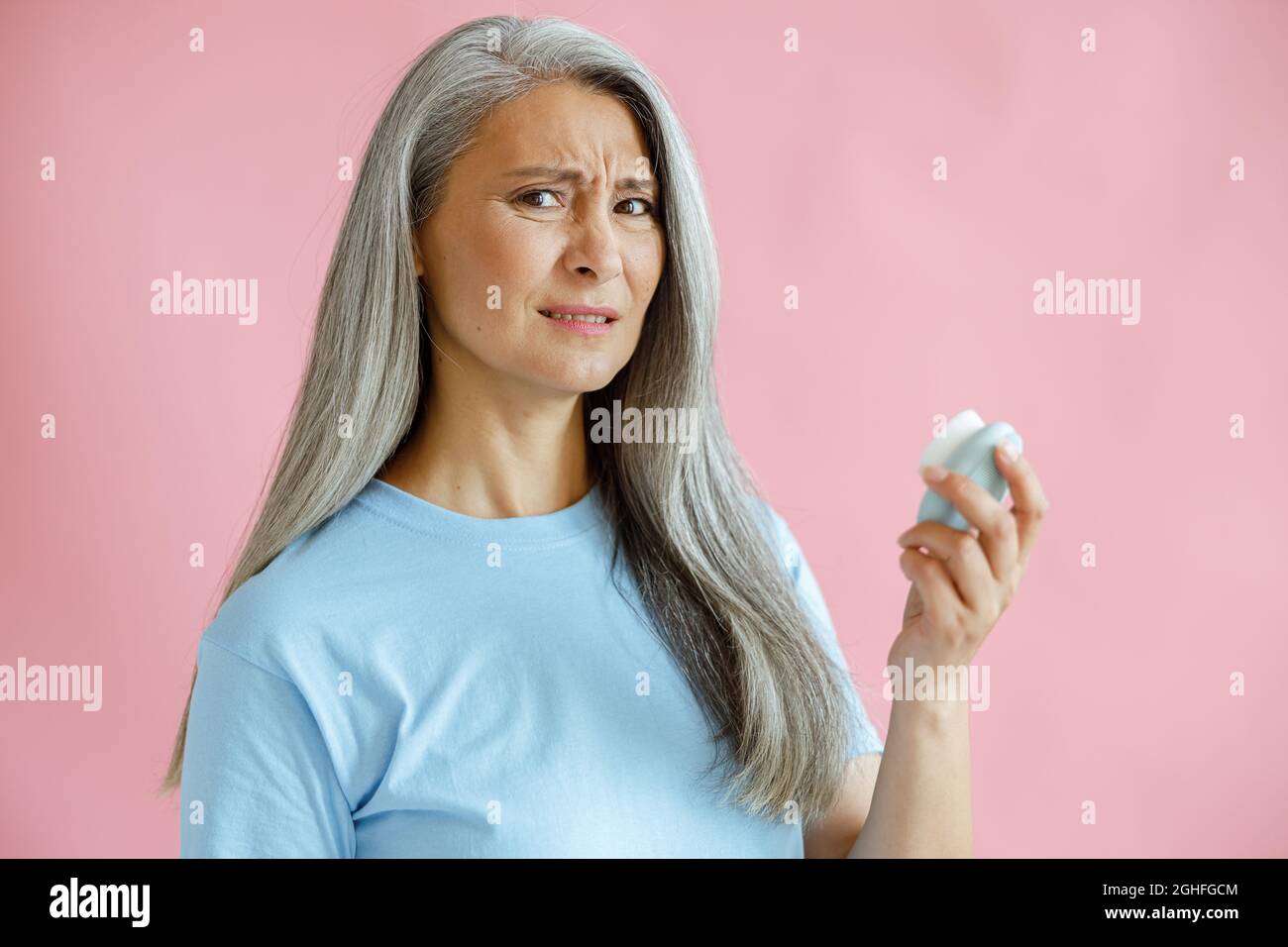 Donna asiatica con capelli grigi disgustosi mantiene la spazzola per la pulizia del viso sonica in studio Foto Stock