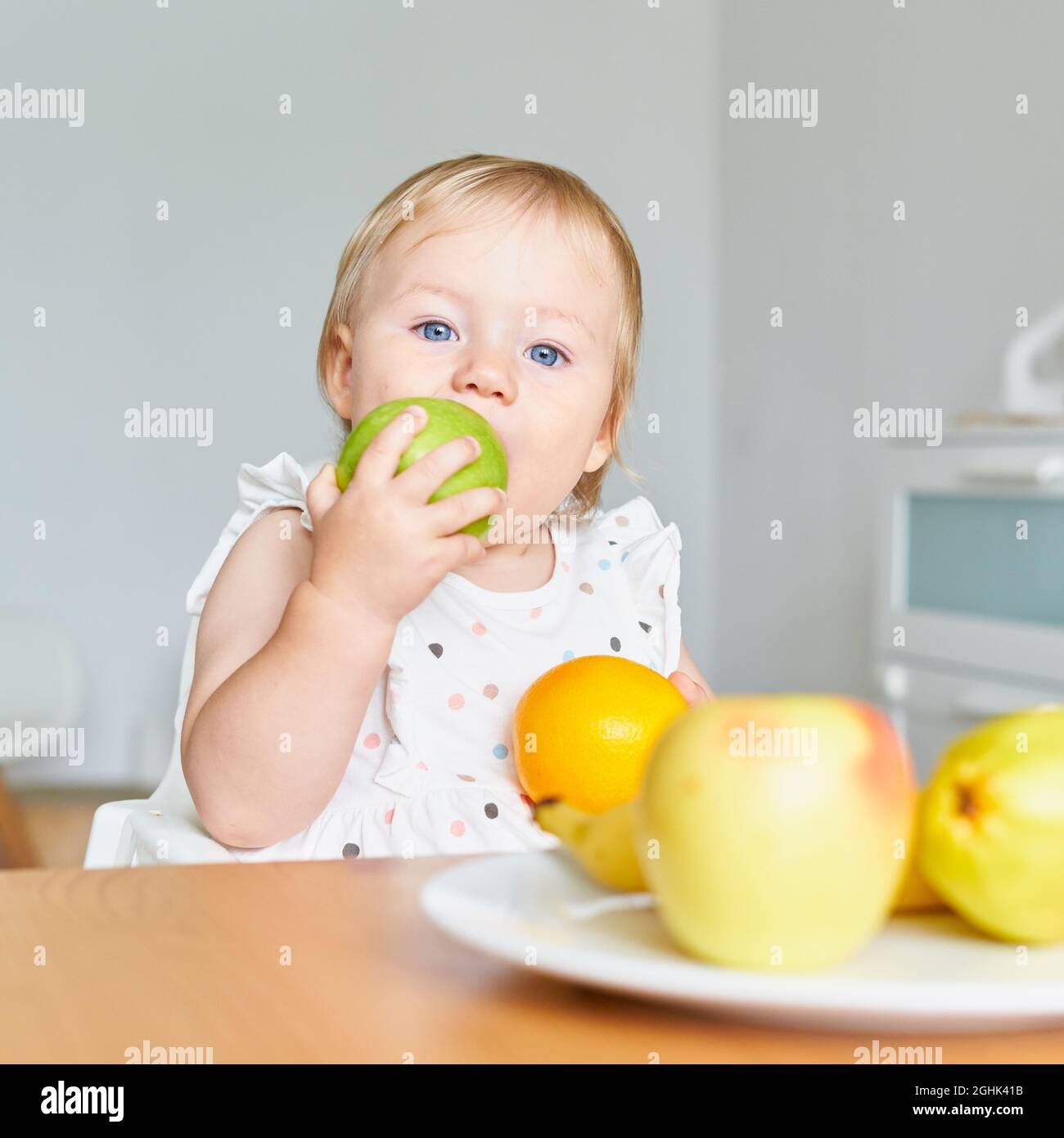 Il bambino biondo con occhi blu mordicchiava l'apla verde e guardava la macchina fotografica. Piatto pieno di frutta Foto Stock
