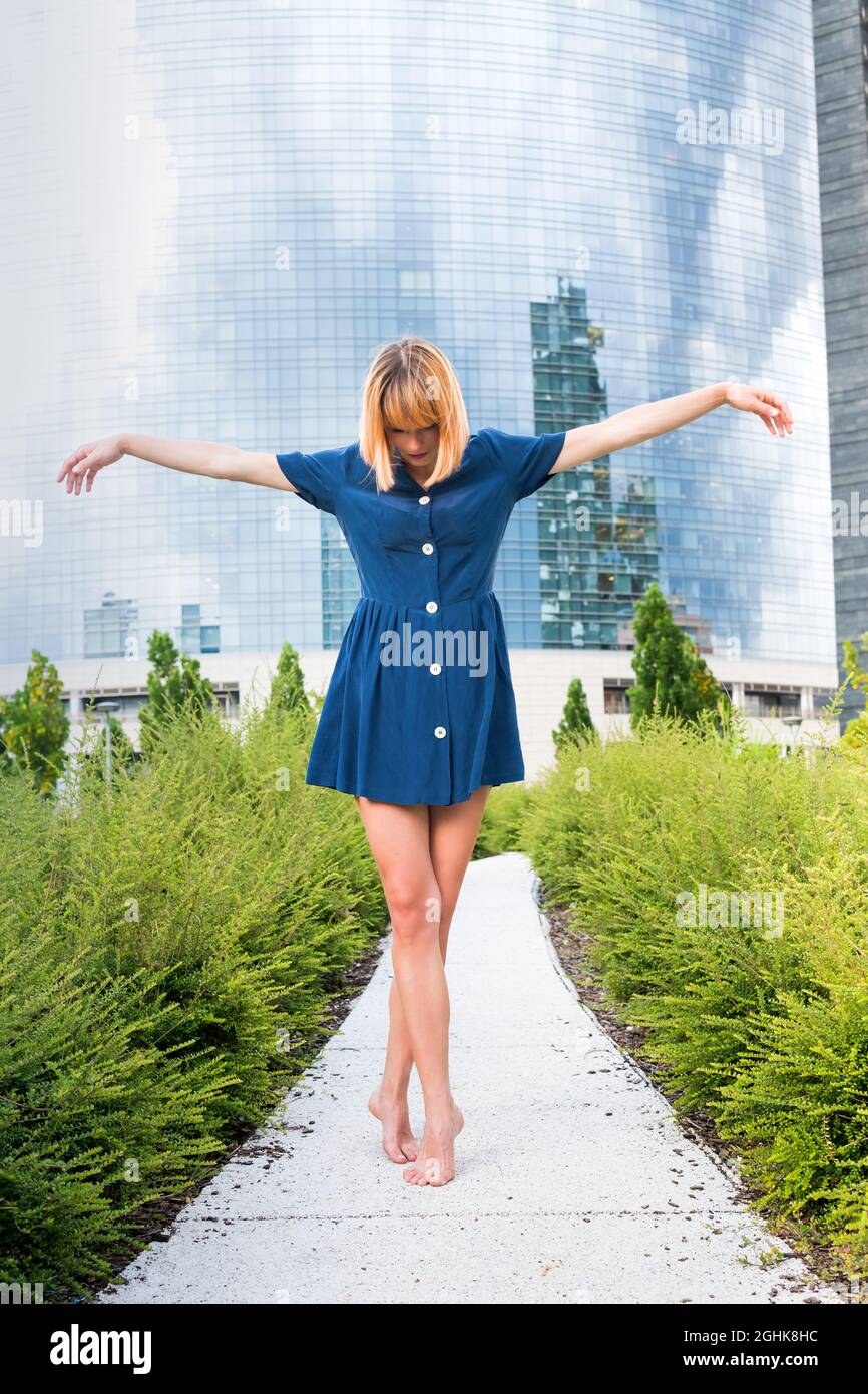 Corpo pieno di giovane donna a piedi nudi in abito blu che balla su punta di lupo con le braccia distese su uno stretto sentiero contro l'edificio urbano contemporaneo con g Foto Stock