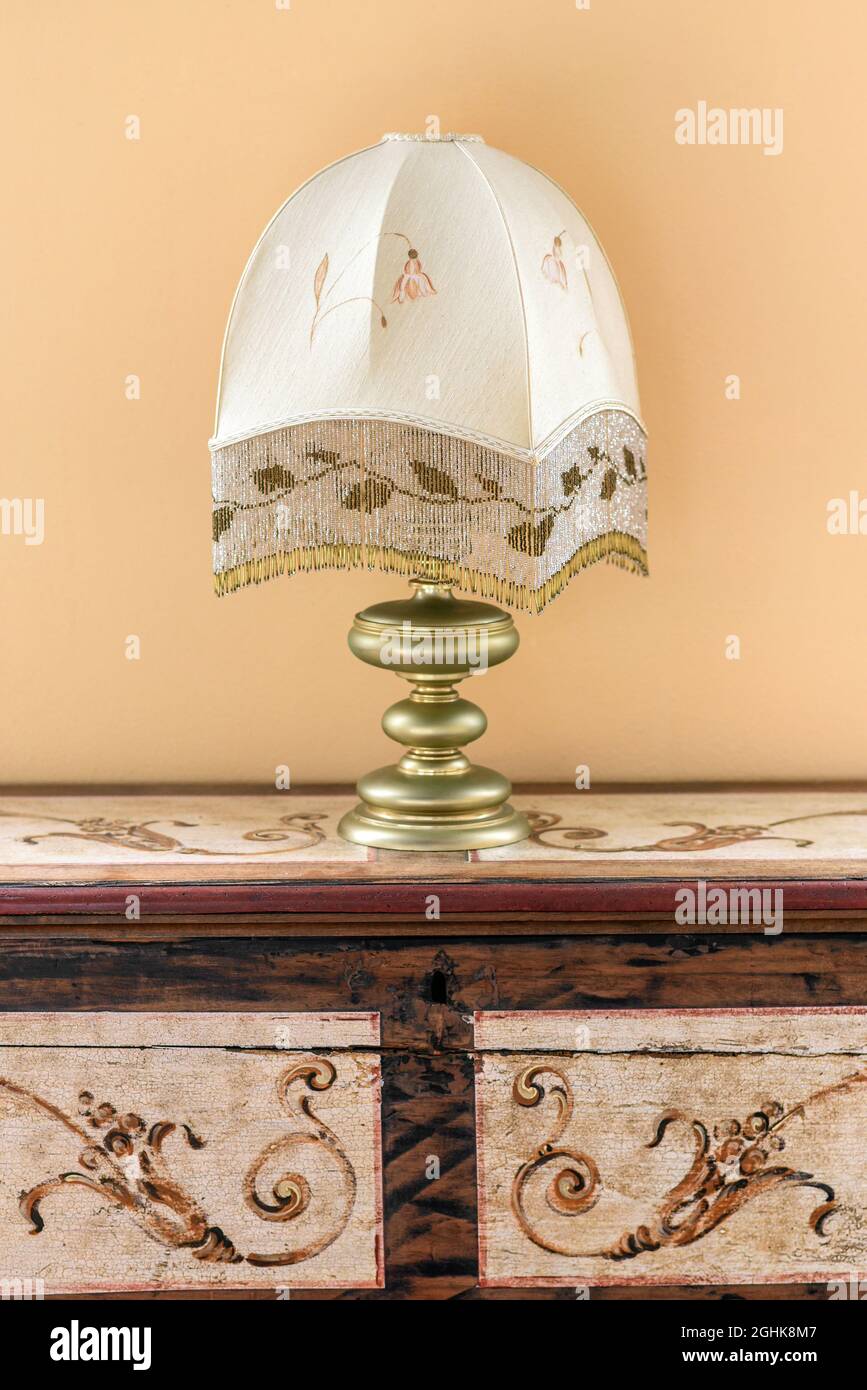 Lampada da tavolo in vecchio stile con paralume a motivi geometrici posta su mobili in legno d'epoca con elementi ornati contro le pareti beige in camera Foto Stock