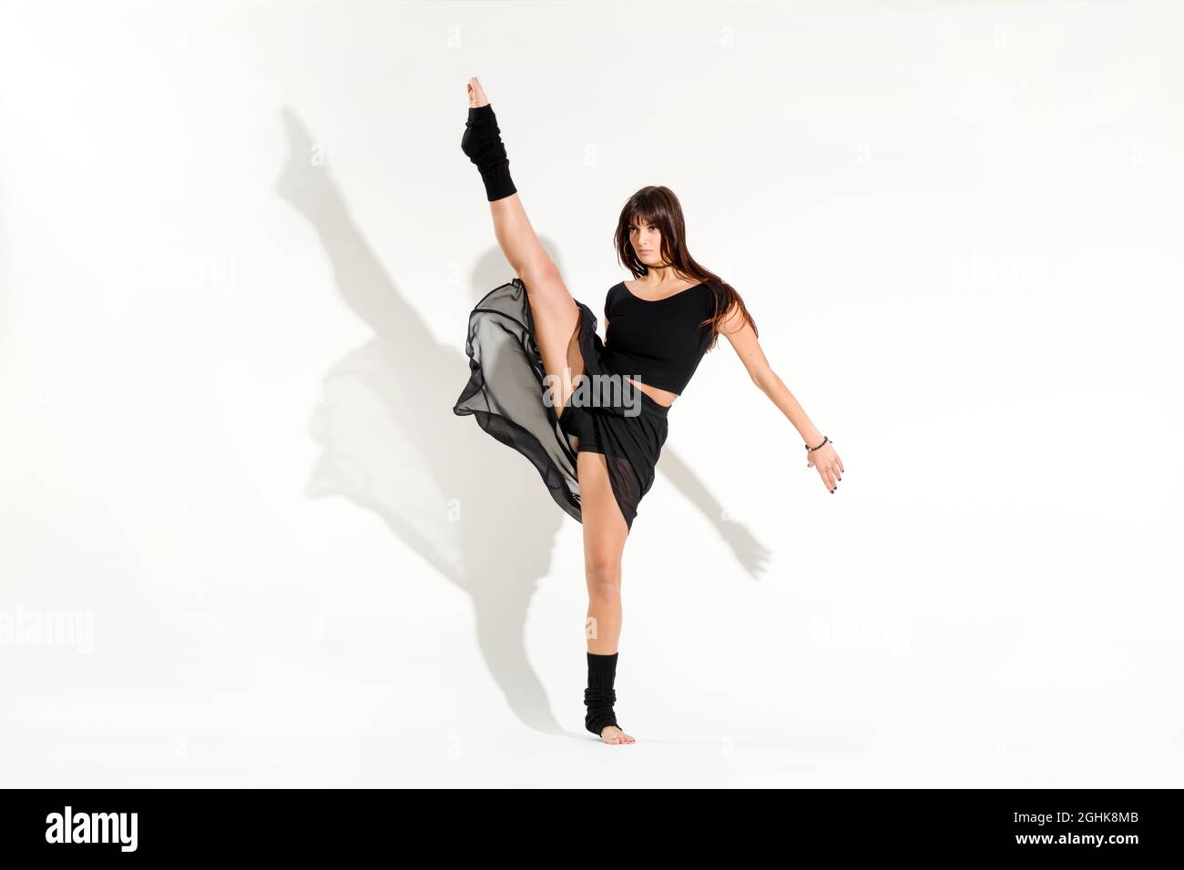 Corpo completo di giovane ballerina a piedi nudi in abito nero che esegue una posa di pattinaggio su ghiaccio dinamica con gamba sollevata contro la parete bianca Foto Stock