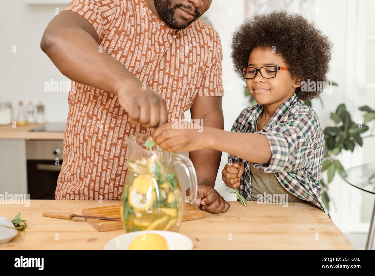 Carino ragazzino che mette foglie di menta secca in caraffa con limonata mescolata dal papà Foto Stock