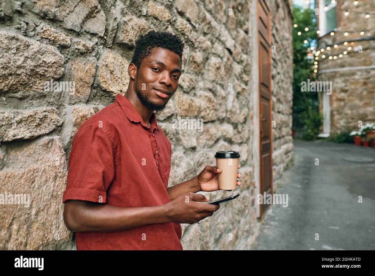 Felice giovane africano in casualwear scorrimento in smartphone e bere mentre in piedi da muro di pietra Foto Stock