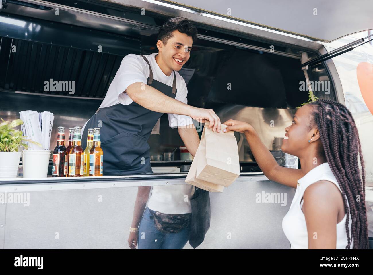 Un venditore sorridente che offre pacchetti con cibo di strada a una cliente femminile. Il proprietario del camion dell'alimento serve l'acquirente. Foto Stock