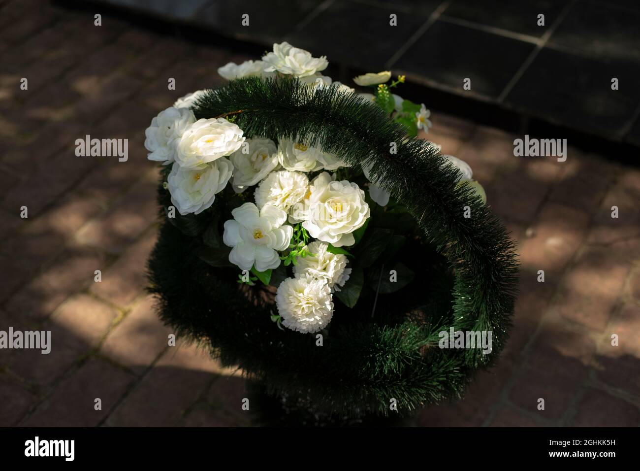 Corona commemorativa di fiori bianchi. Cestino con fiori. Piante artificiali alla luce del sole. Foto Stock