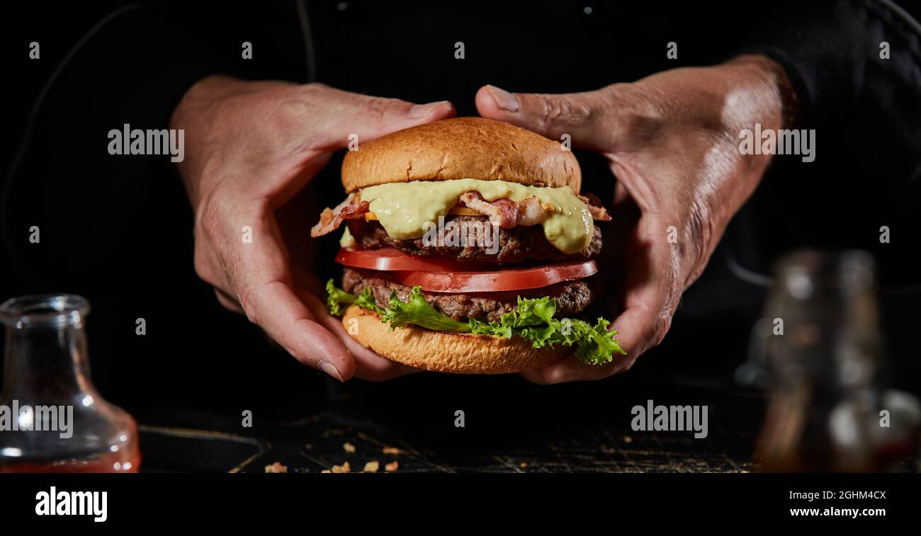 Uomo che tiene un delizioso doppio hamburger di manzo con salsa di senape, pancetta, ingredienti di insalata e succulenti polpette di terra su una boccetta tostata per menu pubblicitari Foto Stock