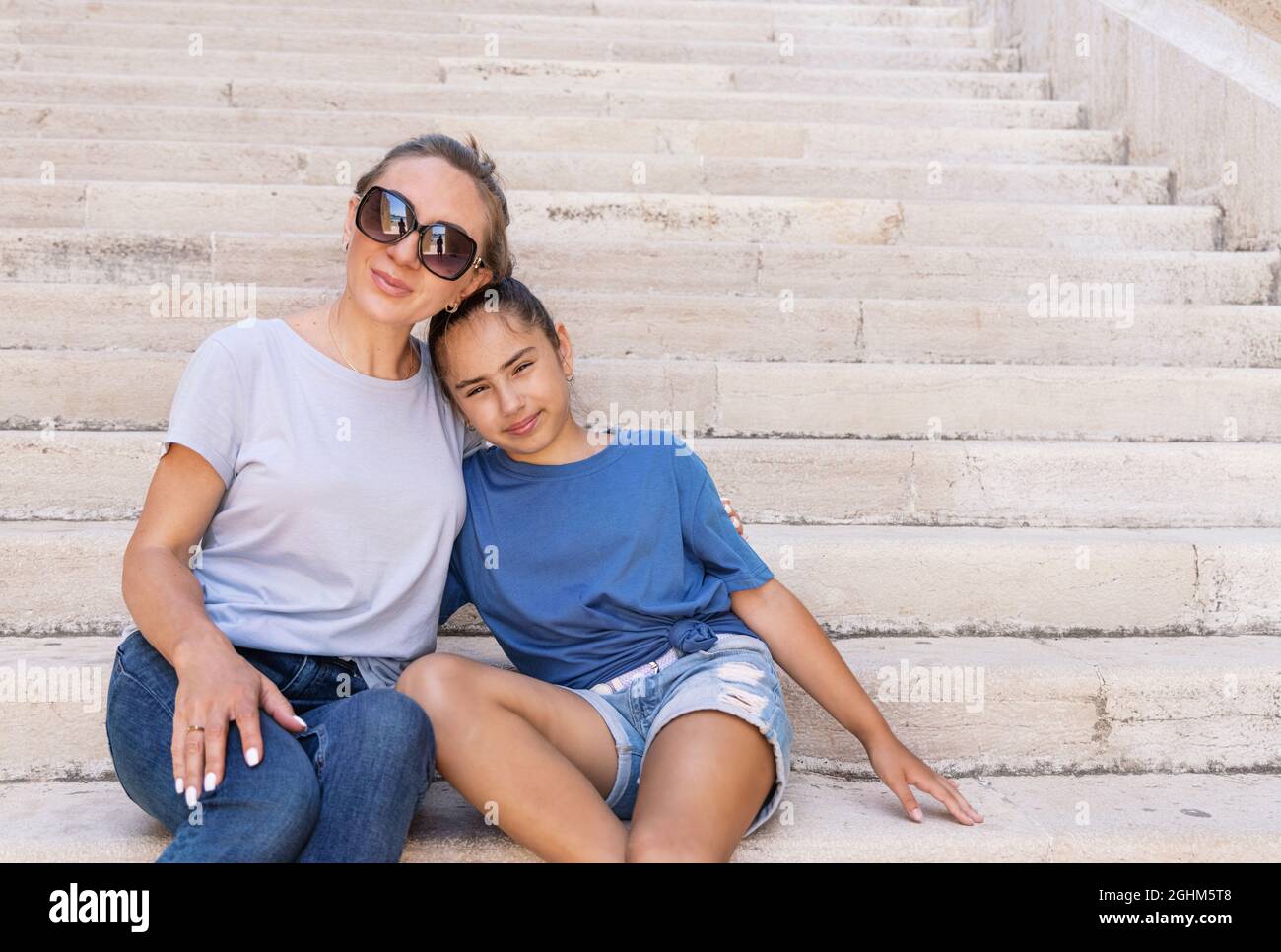 Madre e figlia seduti su scale di pietra all'aperto con t-shirt. Una donna di mezza età e una bambina scolata si abbracciano strettamente. Foto Stock