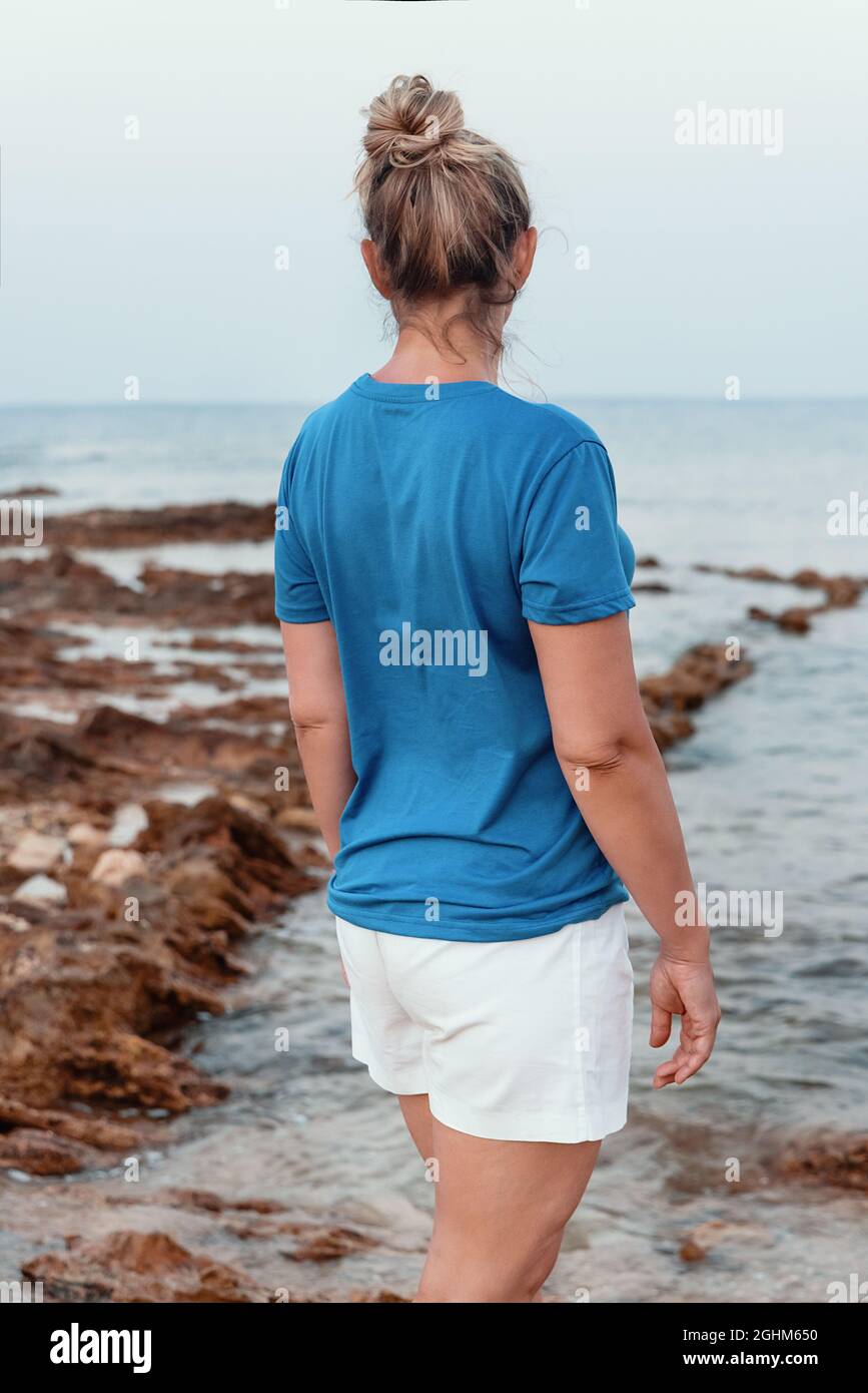 Donne di mezza età in t-shirt blu con bun di capelli in piedi sulla scogliera vicino al mare al tramonto. Back shoot, mockup maglietta Foto Stock