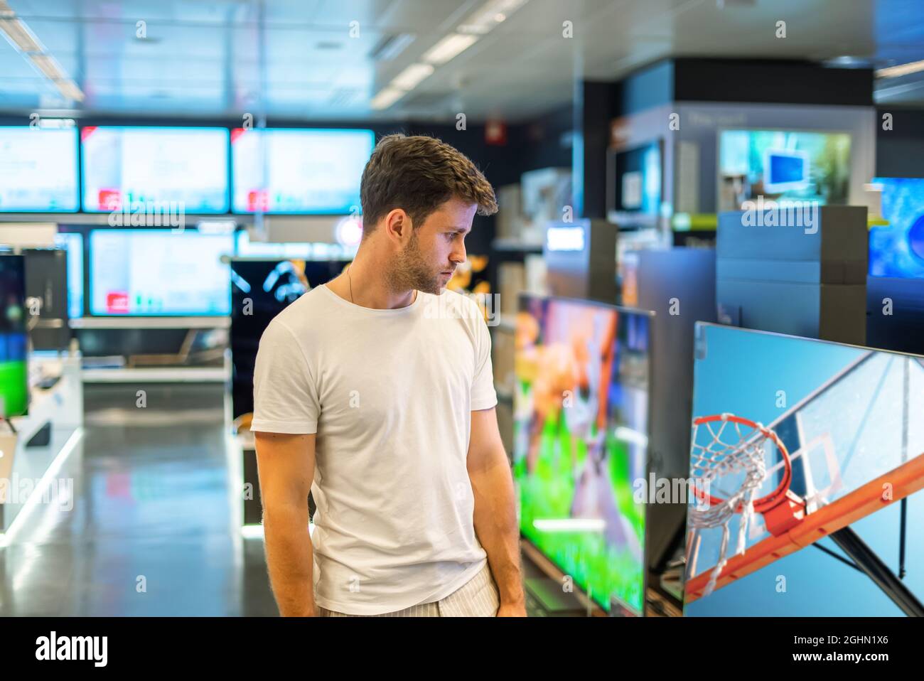 Interessato giovane acquirente maschio guardando moderno Smart TV a schermo piatto con alta risoluzione, mentre la scelta di prodotti in negozio di elettronica spaziosa Foto Stock