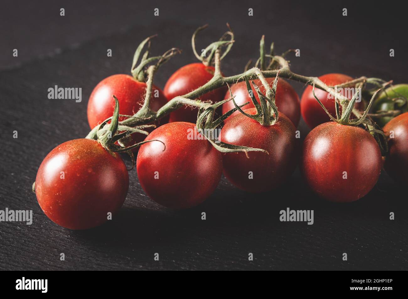 un mazzo di pomodori ciliegini freschi su un ramo si trova su un bordo nero da servire su uno sfondo scuro Foto Stock