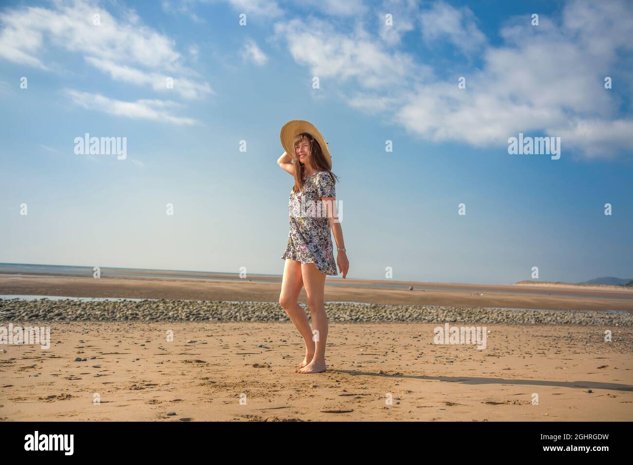 Donna isolata in abito floreale a piedi sulla spiaggia del Regno Unito in vacanza estiva tenendo al suo grande, cappello di paglia sole. Foto Stock