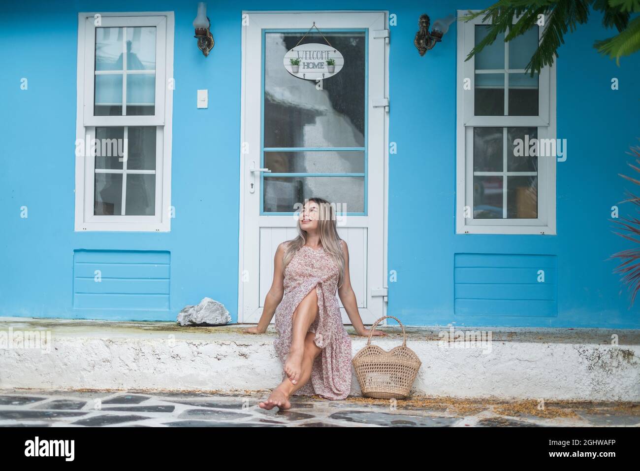 Graziosa giovane donna caucasica con capelli biondi seduti vicino a un'accogliente caffetteria con pareti blu. Bella foto d'estate. Sacchetto di paglia intrecciato Foto Stock