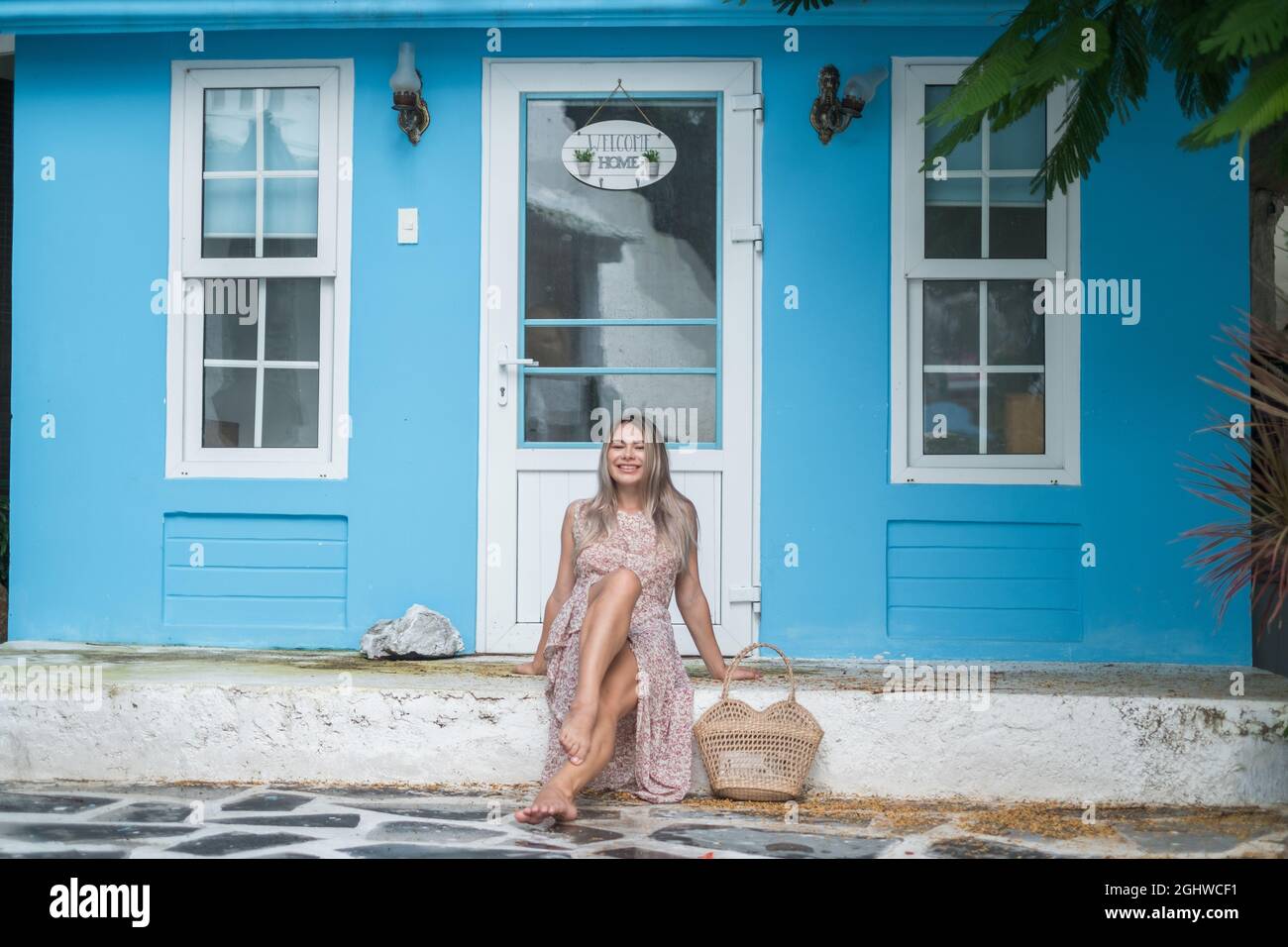 Graziosa giovane donna caucasica con capelli biondi seduti vicino a un'accogliente caffetteria con pareti blu e sorridenti con i denti. Ragazza ridente. Sacchetto di paglia intrecciato Foto Stock