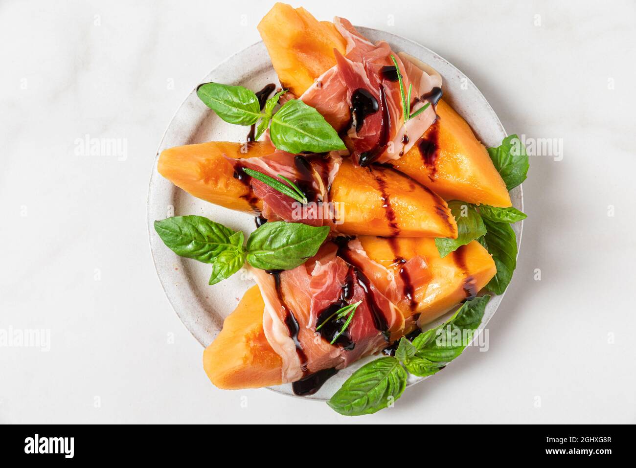 Melone cantalupo fettine con prosciutto, salsa all'aceto balsamico e basilico in un piatto su sfondo bianco. Antipasto italiano. Vista dall'alto. Foo sano Foto Stock