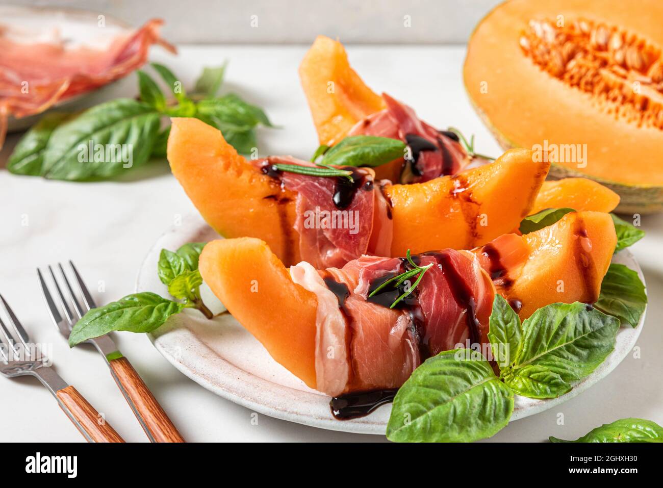 Prosciutto con fette di melone cantalupo, salsa all'aceto balsamico e basilico in un piatto con forchette su fondo bianco. Antipasto italiano. Primo piano. Foto Stock