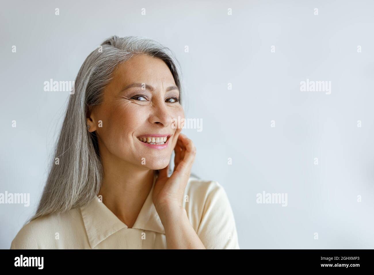 Donna dai capelli argenti in camicia beige che tocca la guancia su sfondo grigio chiaro Foto Stock