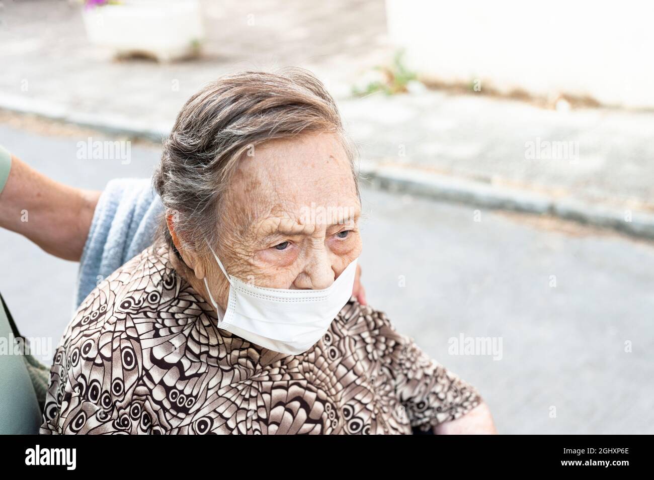 primo piano di una donna ottogenaria che cammina lungo la strada in una sedia a rotelle, rivolta in avanti in una camicia a motivi geometrici, indossando una maschera protettiva. Foto Stock