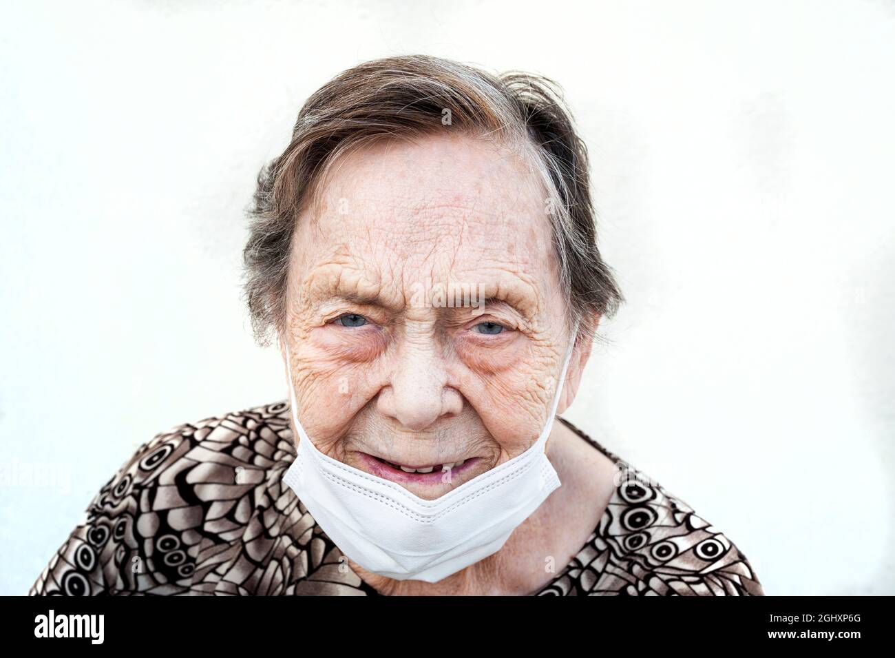 Primo piano di Donna di oltre 80 anni sorridente con maschera protettiva giù. Faccia con le rughe dell'età e gli occhi blu-grigi. Indossare una camicia stampata. Bianco ba Foto Stock