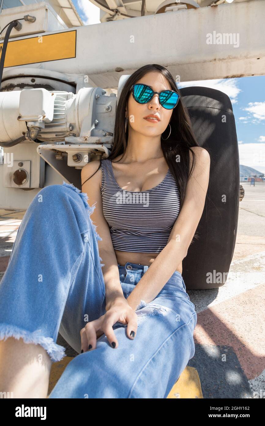 riposandosi all'ombra accanto ad un pneumatico una bella giovane donna  latina con capelli lisci neri, occhiali da sole accessori moda, top grigio  e jeans, urban Foto stock - Alamy