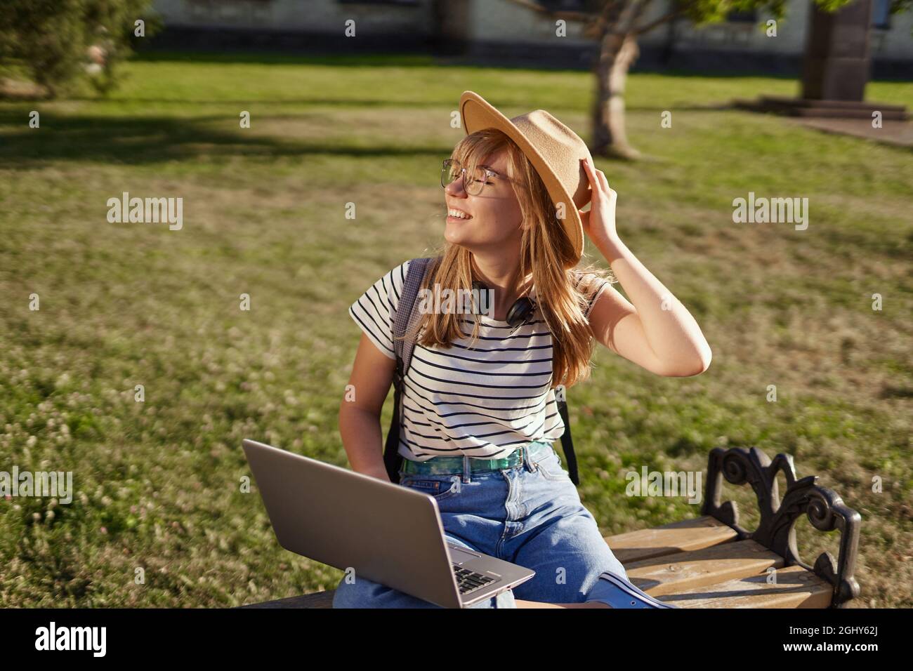 Allegro e sorridente università o studente universitario con zaino, occhiali e cappello utilizzando un computer portatile seduto sul banco del campus universitario. Concetto di homeschooling, e-learning, istruzione o surf Foto Stock