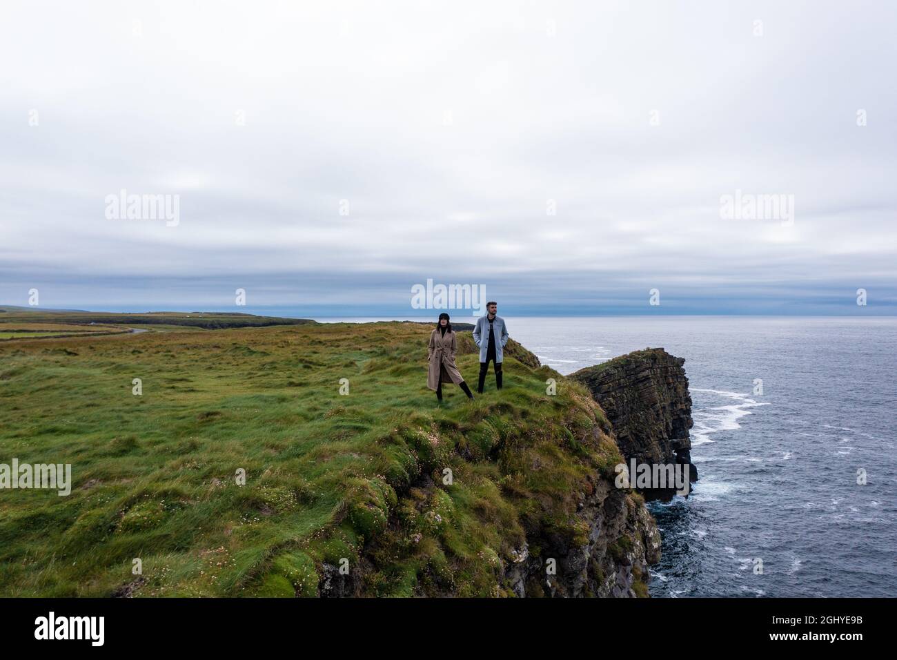 Giovane uomo e donna che indossa una giacca in piedi ai margini della scogliera di Moher in Irlanda mentre viaggia e goditi la natura durante il giorno sotto il cielo nuvoloso Foto Stock