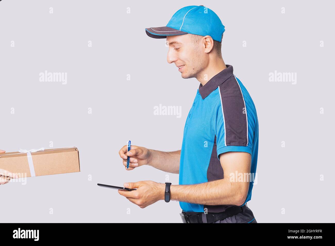 Vista laterale foto di un uomo di consegna in uniforme postman indossare orologio digitale sulla sua mano e tenere tablet con una penna per firmare per la confezione. Foto Stock