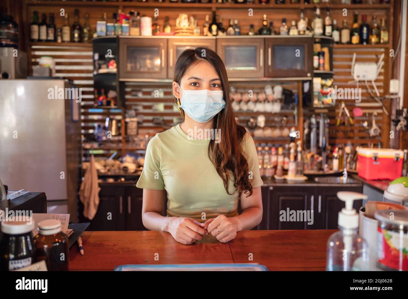 Attraente giovane imprenditore asiatico donna indossare maschera in piedi per accogliere i clienti al banco in legno nel ristorante Foto Stock
