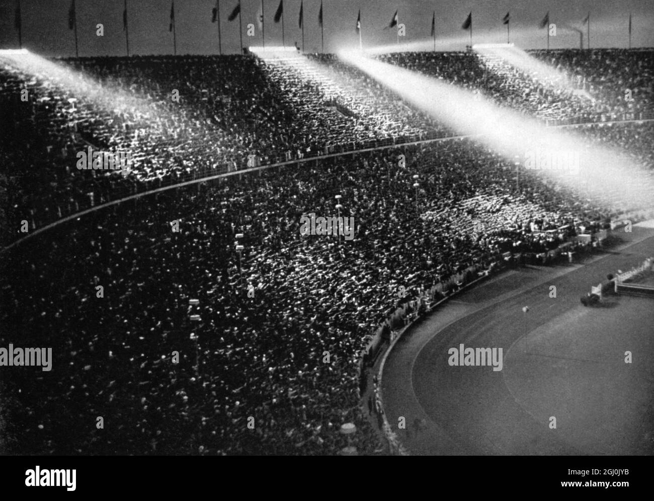 1936 Olimpiadi, Berlino - i riflettori tagliano la notte e si depositano sulle teste della massa di spettatori qui sotto. (Die Scheinwerfer jagen Lichtbundel uber die Menschenmassen, Fanfaren schneiden wie Schwerter die Bander der Nacht.) ©TopFoto Foto Stock