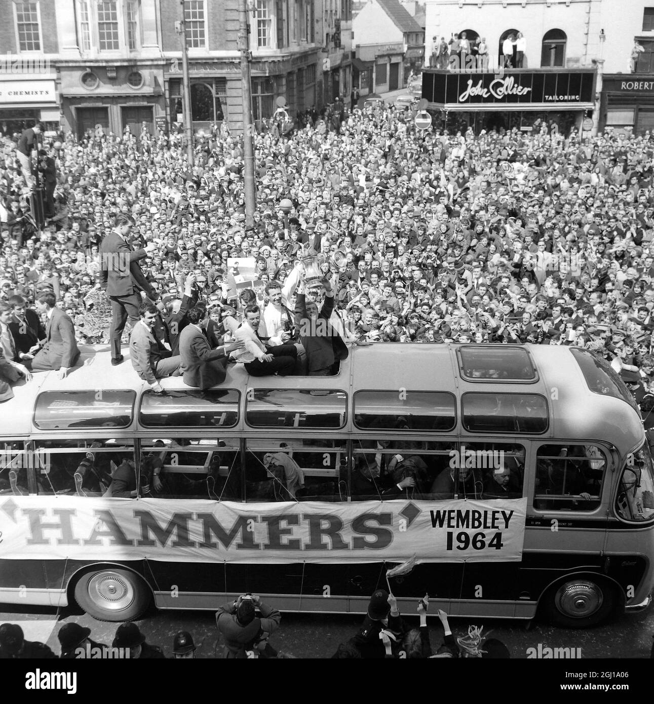 3 MAGGIO 1964 Una sezione delle migliaia di Eastenders costeggiano la strada a Stratford Broadway nell'East End di Londra come West Ham United Football Team, vincitori della finale della fa Cup di ieri a Wembley, sfilò per le strade di West e East Ham con l'ambito trofeo. Con orgoglio che sfoggia il trofeo è il capitano del prosciutto occidentale Bobby Moore. Dietro di lui ci sono Ken Brown (a destra) e Johnny Byrne (terzo da destra). Così tante persone si sono affollate nel municipio di Stratford che il sindaco ha dovuto chiamare fuori la reception ufficiale per il team. La polizia era preoccupata che se la squadra una volta lasciasse il loro allenatore, non sarebbero stati in grado Foto Stock