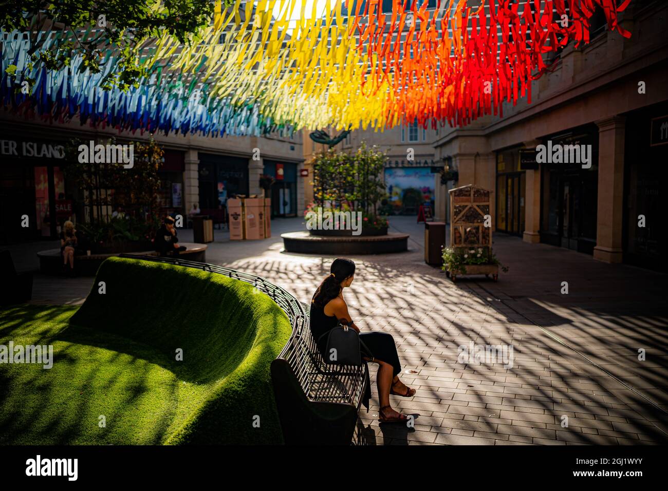 Una persona si siede sotto le decorazioni di strada color arcobaleno mentre  il forte sole del mattino proietta ombre sul pavimento nel centro di Bath.  Data foto: Mercoledì 8 settembre 2021 Foto
