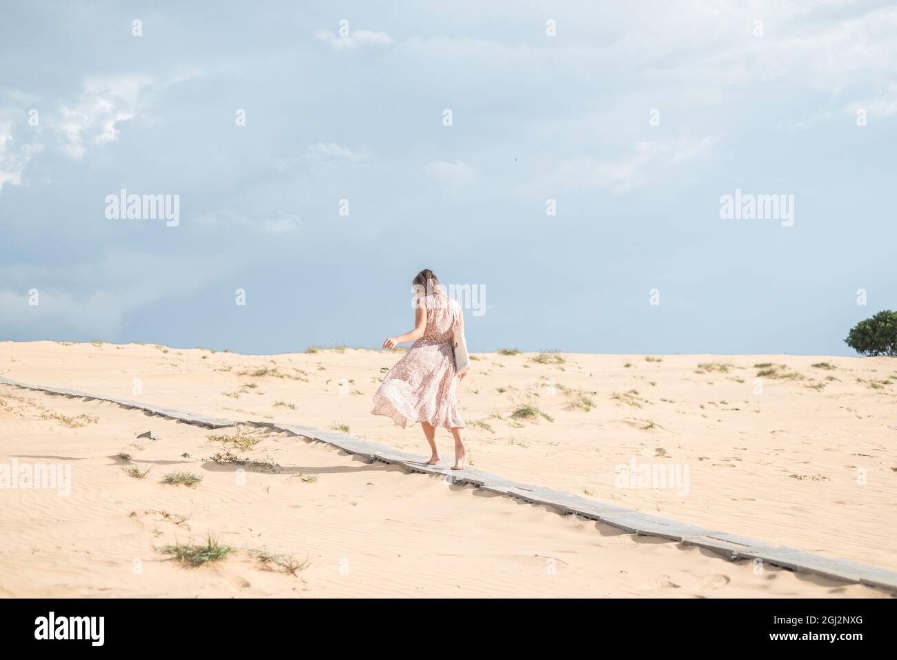 Bella vista della donna bionda caucasica in abito lungo a piedi nel deserto con sabbia bianca in giorno di sole durante le vacanze estive. Dune di sabbia. Foto Stock