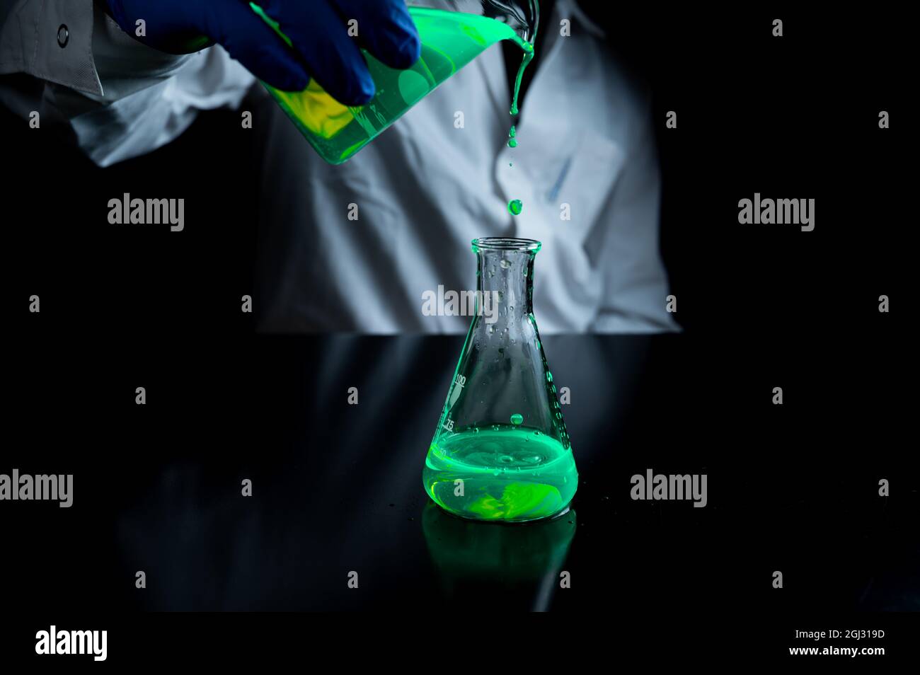 Uno scienziato di ricerca che sperimenta con gocce fluorescenti verdi in un pallone conico di vetro in laboratorio biomedico scuro per medicina di cura della salute d Foto Stock