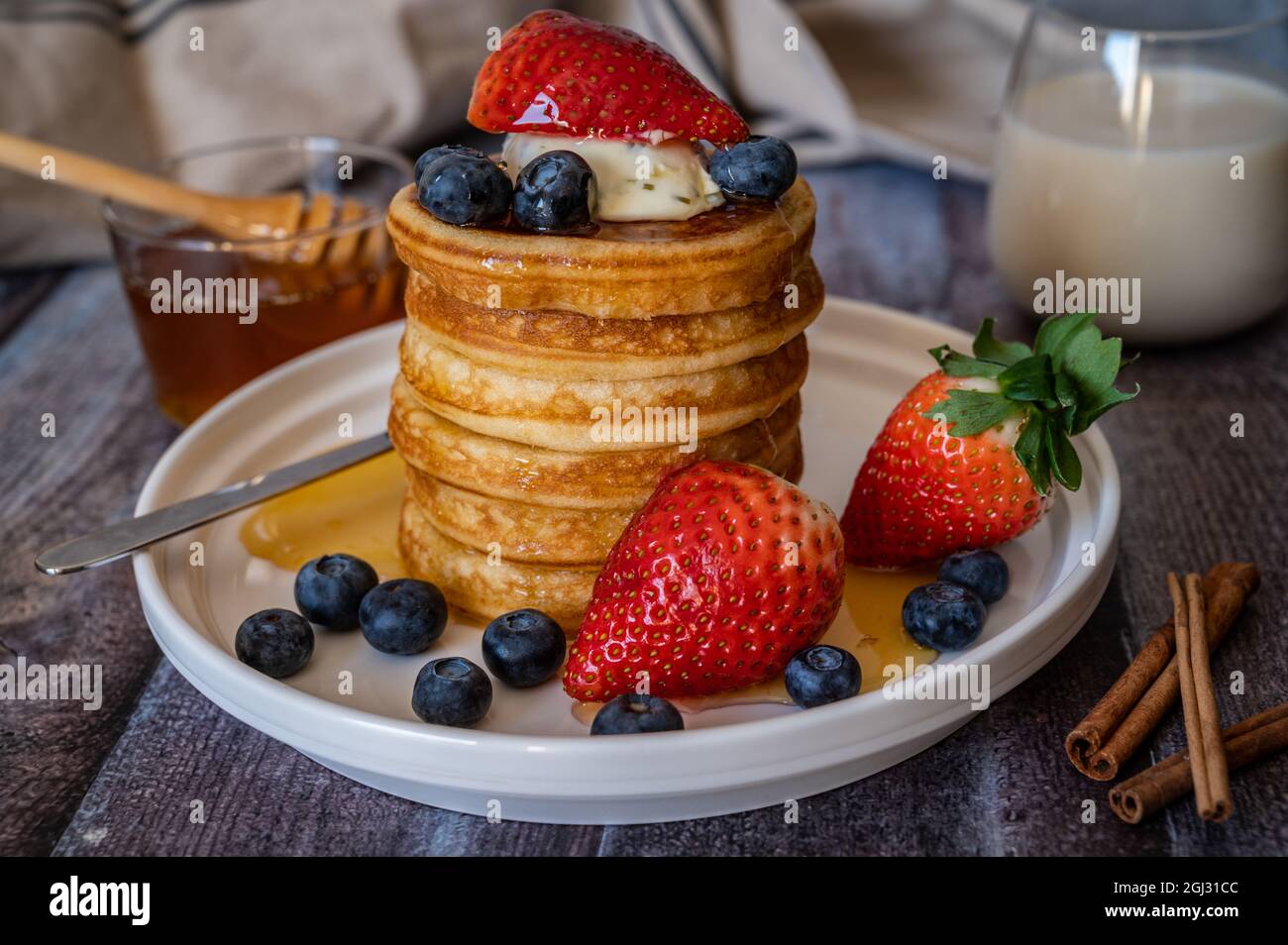 Vista in primo piano su una pila di pancake con bastone di miele, fragola, bacca blu e panna a frusta in cima al piatto, delizioso dessert con latte per colazione. Foto Stock