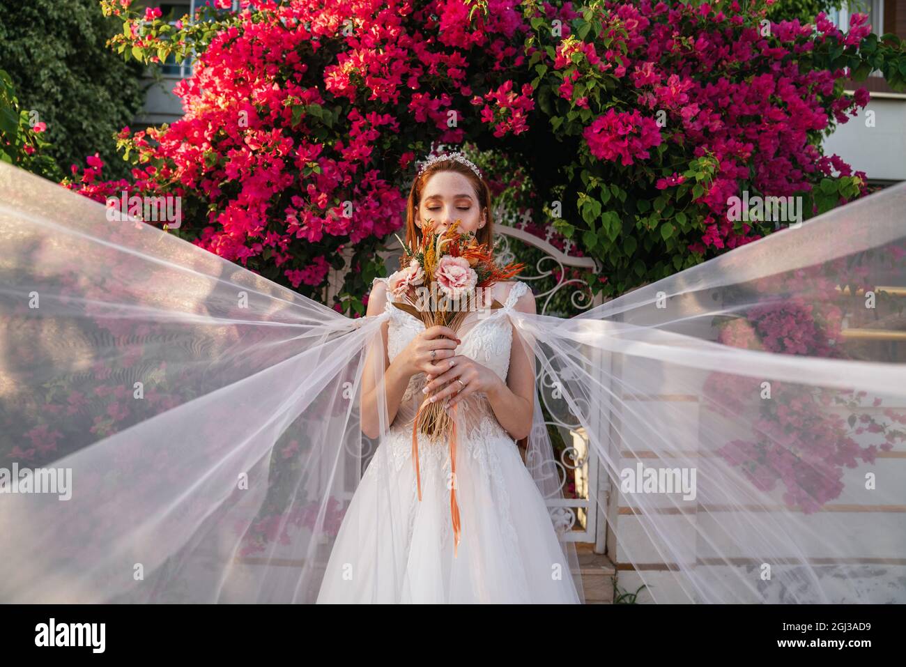 Sposa con abito da sposa bianco che tiene bouquet colorato e secco per matrimoni con fondo rosso buganvillea. . Foto di alta qualità Foto Stock