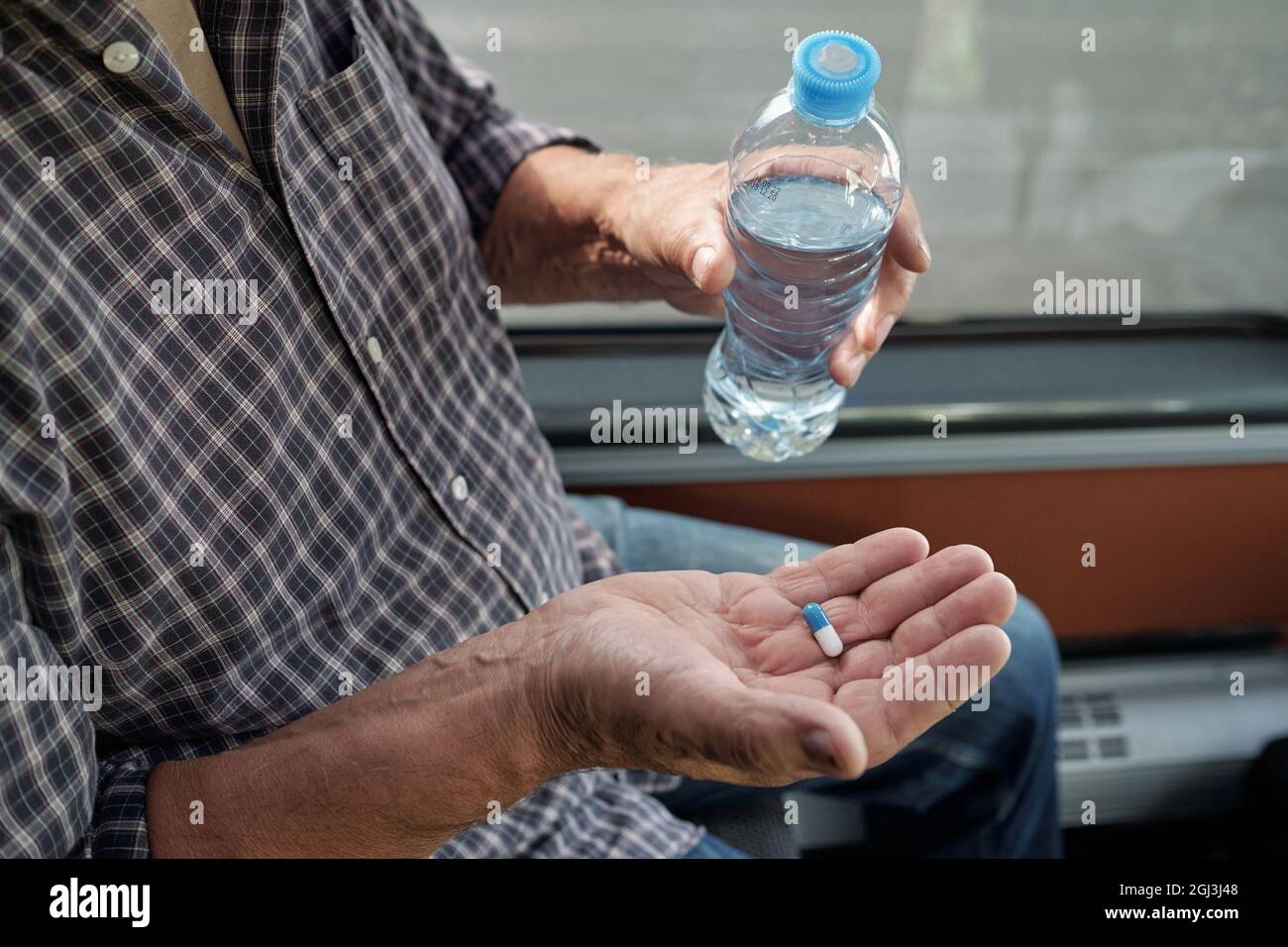 Primo piano di un uomo anziano irriconoscibile in una camicia casual che tiene una bottiglia d'acqua e prende la pillola di dramamine in autobus Foto Stock
