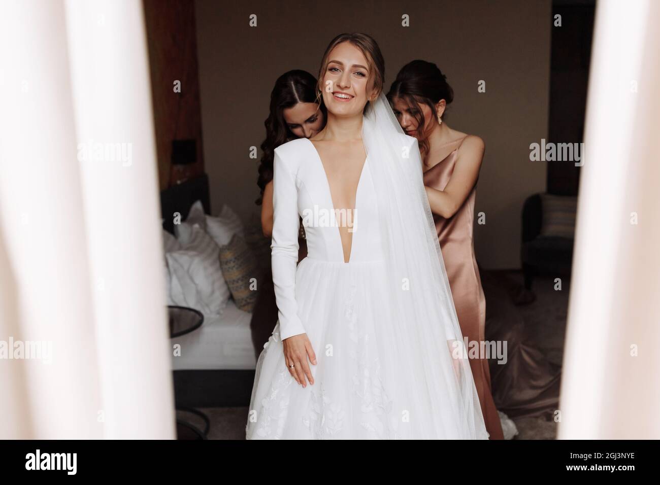 bella giovane sposa sorridente in lungo vestito bianco satin con profondo  scollo, acconciatura e make up in piedi in camera d'albergo. bridesmaids  aiutare la sua preparazione Foto stock - Alamy
