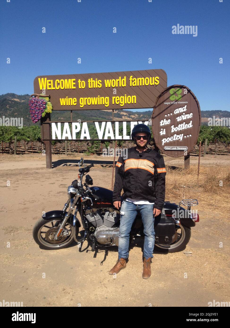 Colpo verticale di un uomo che si appoggia su una moto di fronte al cartello d'ingresso della Napa Valley a Oakville Foto Stock