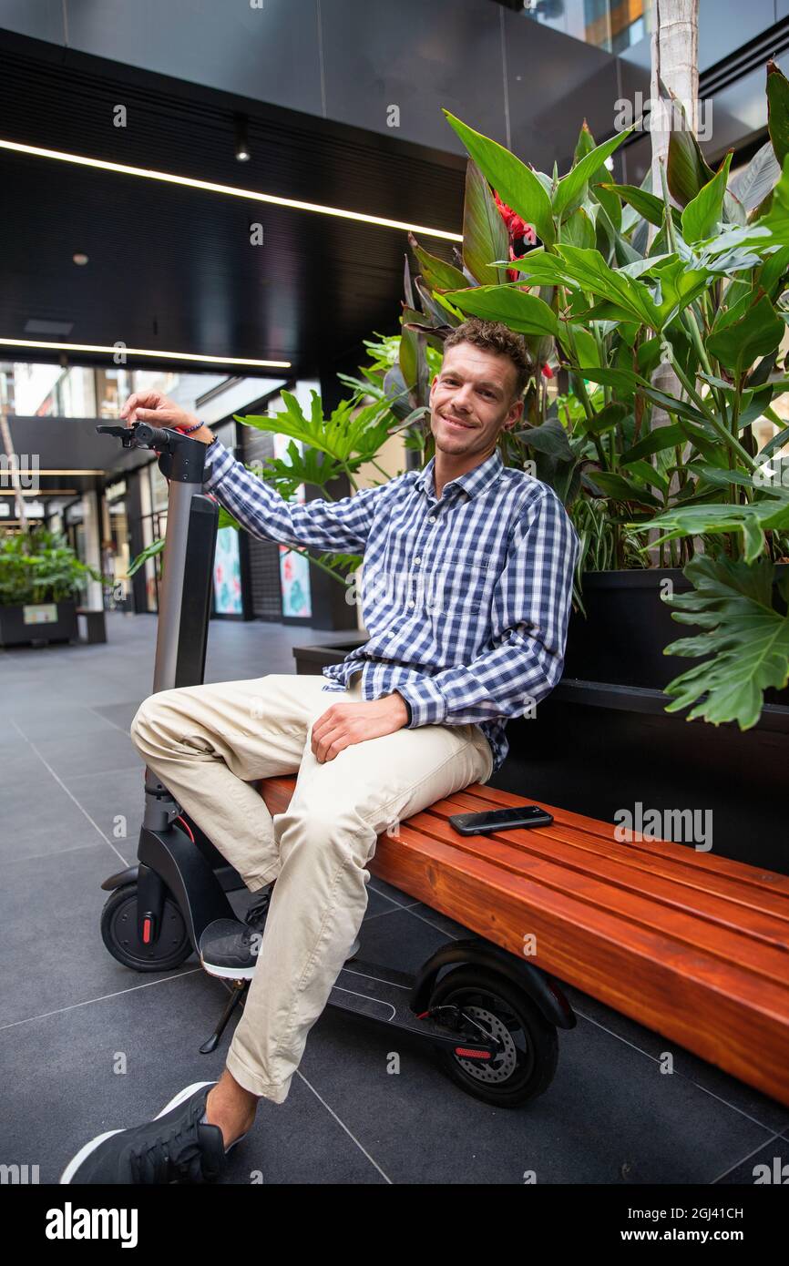 Uomo millenario con il suo scooter elettrico seduto in panchina con il suo smartphone. Ragazzo caucasico ben vestito - concetto di mobilità ambientale-amichevole Foto Stock