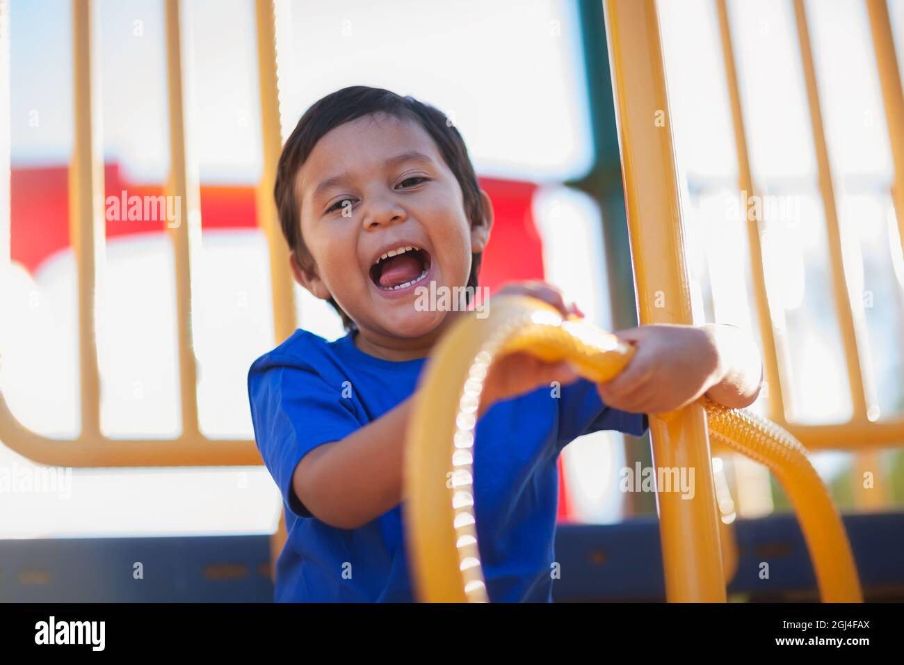 Ragazzo latino che esprime gioia e ridere mentre si tiene in mano le barre di arrampicata su un parco giochi per bambini o palestra di arrampicata all'aperto. Foto Stock