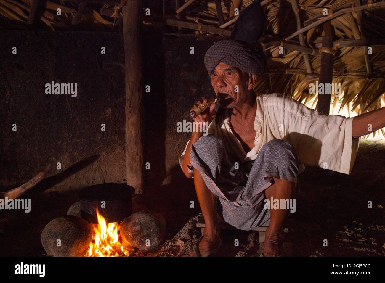 Uomo birmano locale fumare sigaro dal fuoco nella sua casa a Bagan, Myanmar. Foto Stock