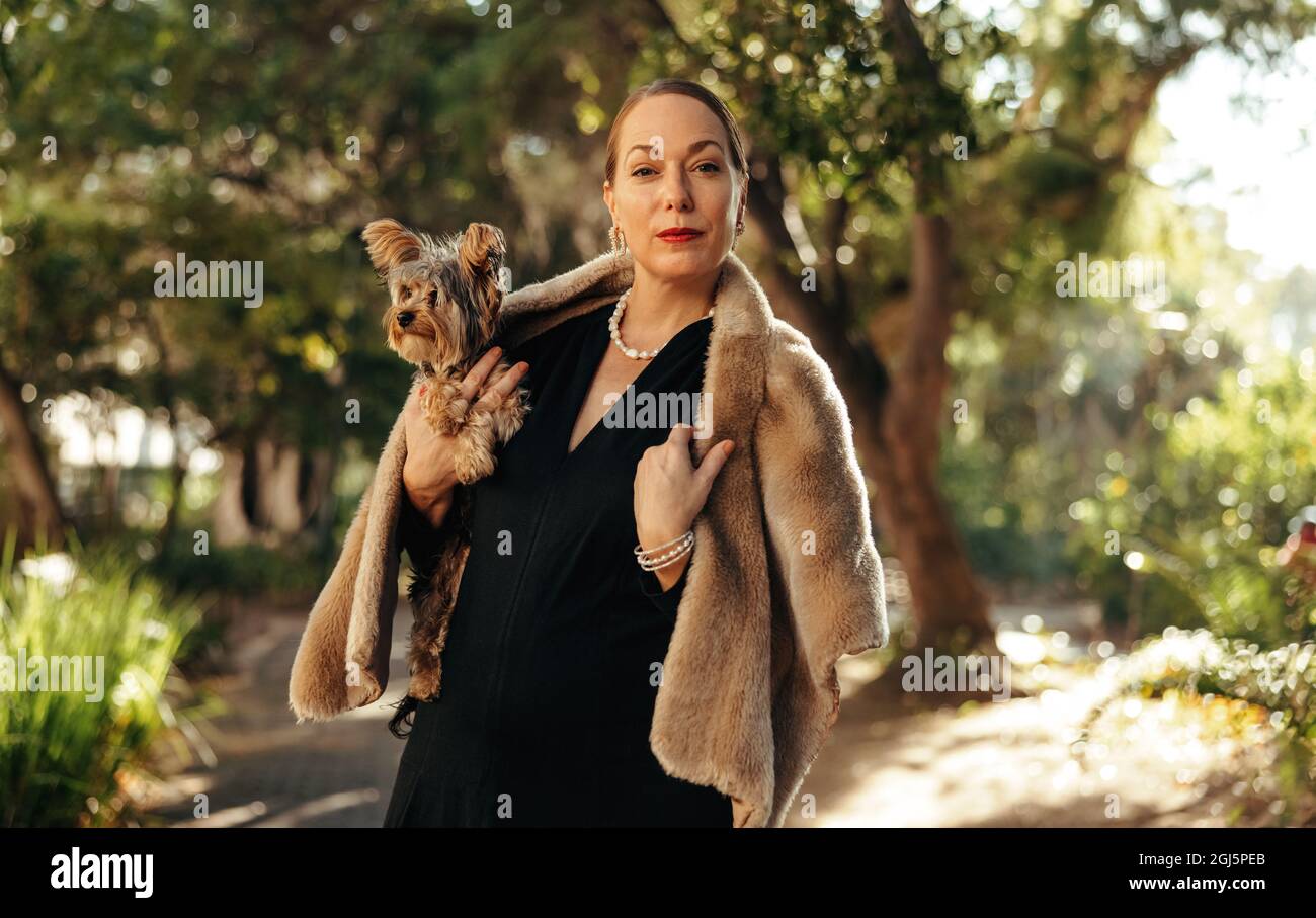 Donna di lusso che guarda la macchina fotografica mentre tiene il suo cucciolo all'aperto. Donna di alta classe in piedi da sola in un parco durante il giorno. Donna animale domestico proprietario goi Foto Stock