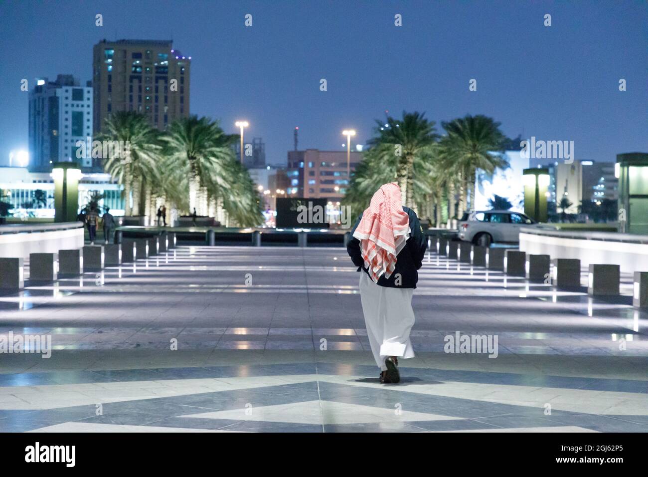 Stato del Qatar, Doha. Uomo vestito tradizionalmente camminando lungo la passerella di marmo la sera. Foto Stock