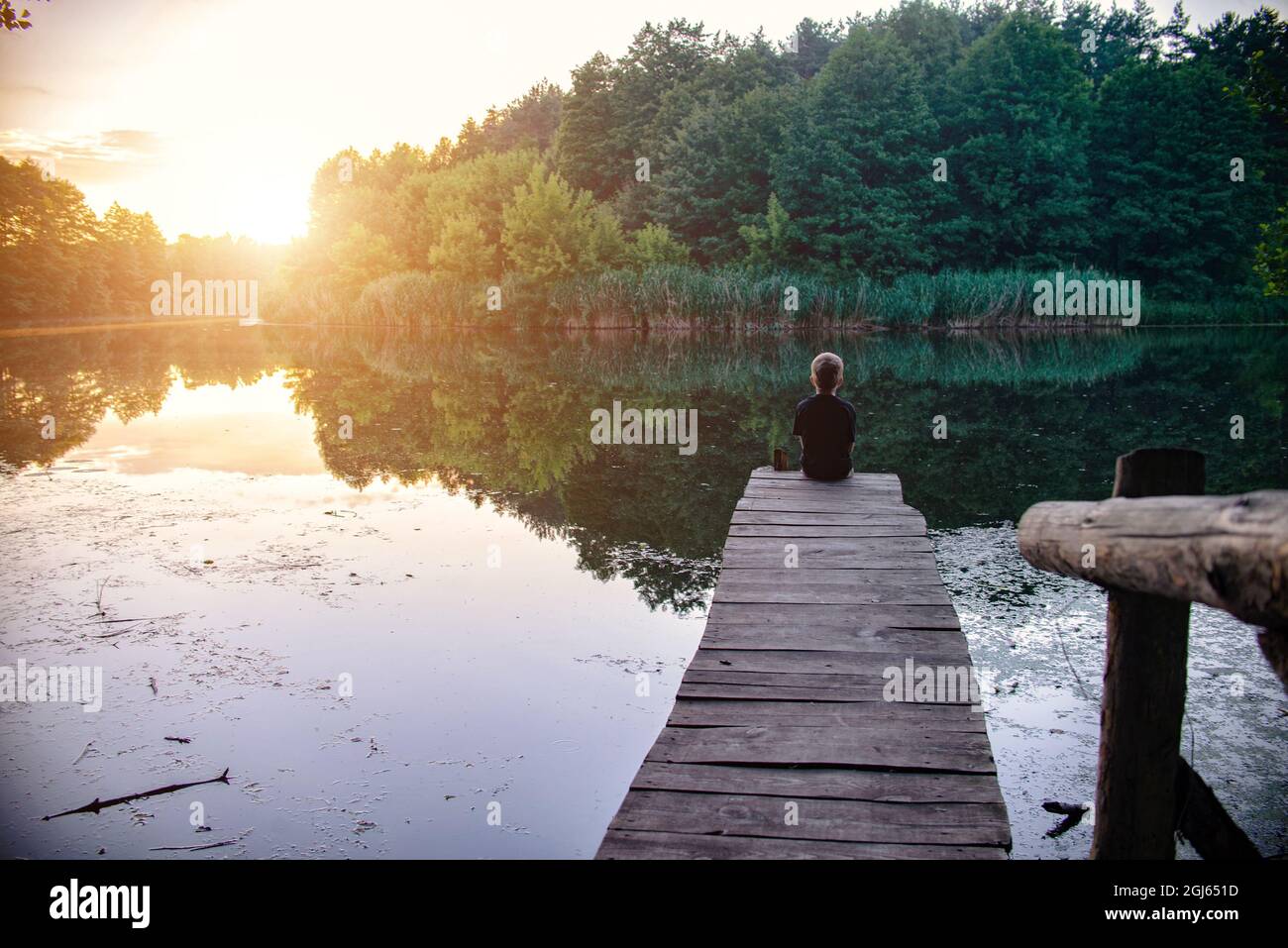 Un ragazzo siede su un ponte di legno sul fiume in un ambiente pittoresco al Sunset.Toning Foto Stock