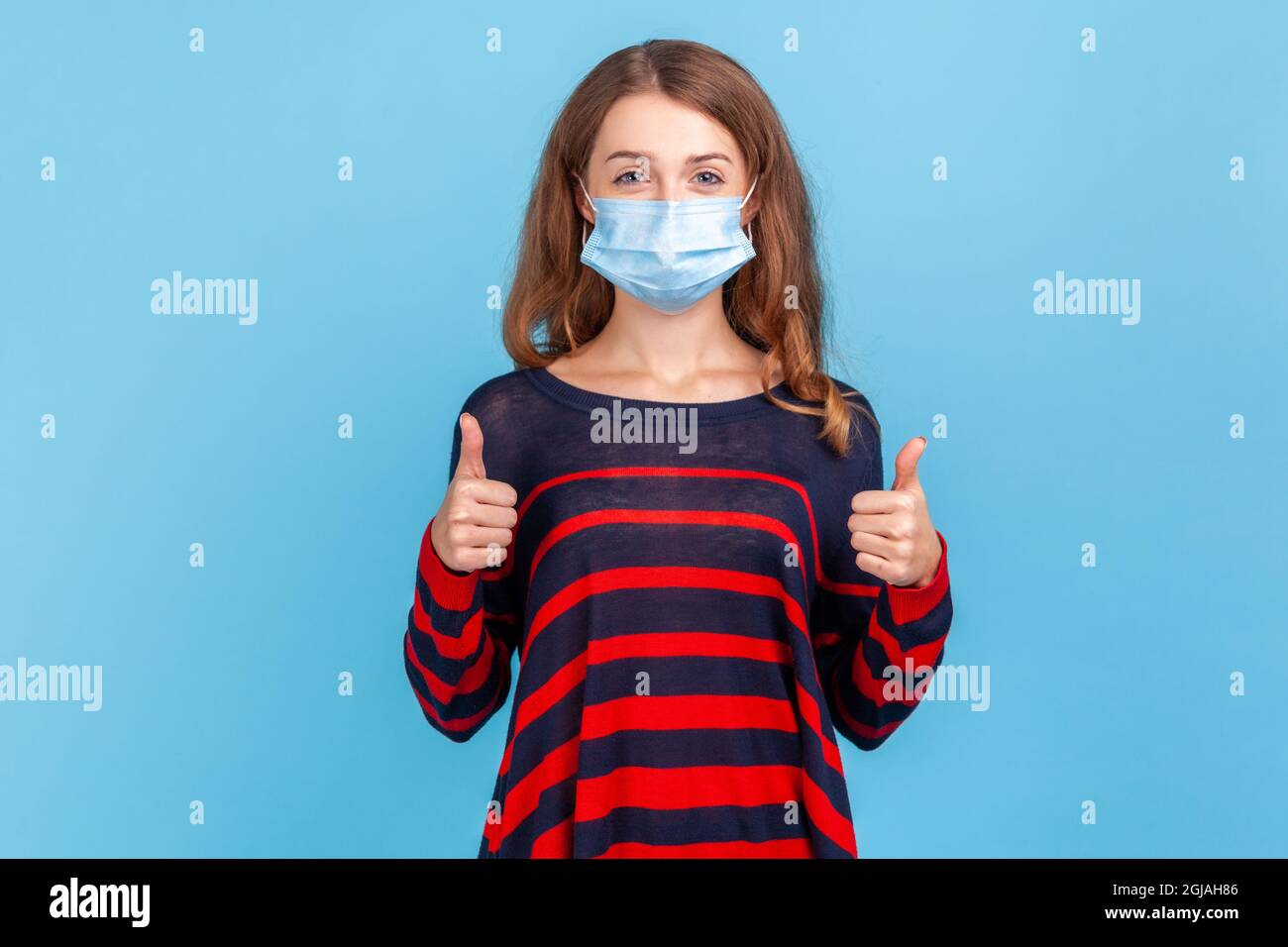Donna che indossa un pullover casual a righe e una maschera medica che mostra il pollice in su, guardando la macchina fotografica, essendo protetto da malattie respiratorie. IO Foto Stock